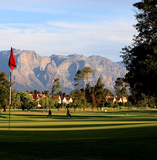 A red flagstick on a green with golfers in front of mountains in Paarl.