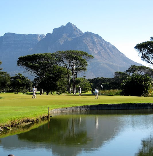 Golfers on a green surrounded by pine trees with Table Mountain in Cape Town.