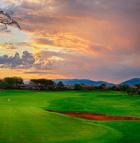 A green and sand bunker with residential homes under a sunset sky near the Magaliesberg mountains.