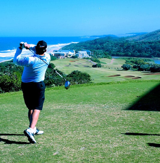 A golfer following through on a drive overlooking fairways and ocean waves on the Dolphin Coast.