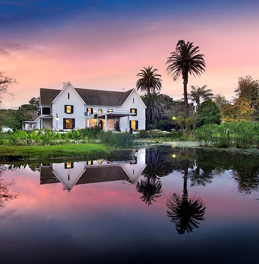Exterior view of a white building surrounded by gardens and palm trees at dusk in the Garden Route, South Africa.