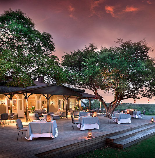 Lantern-lit outdoor dining deck under a large tree at dusk.