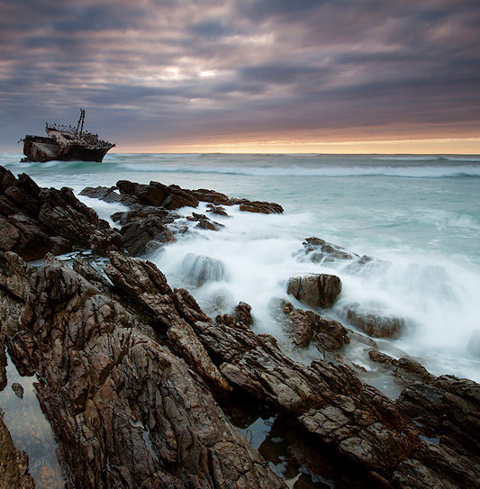 Waves crashing over jagged rocks near a shipwreck at dusk in Agulhas National Park.