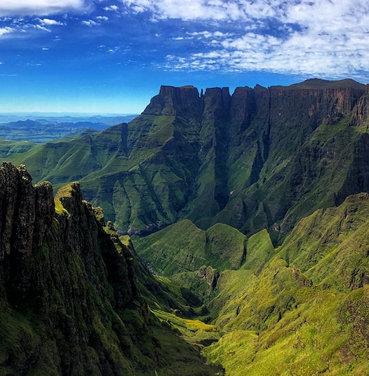 Sunlight illuminating the massive green cliffs and deep mountain valleys of uKhahlamba-Drakensberg National Park under a blue sky.