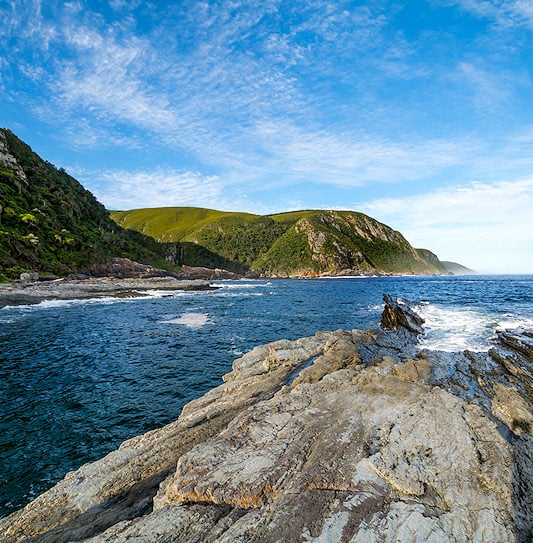 Jagged rock formations extending into the ocean along the steep, forested coastline of Tsitsikamma National Park.