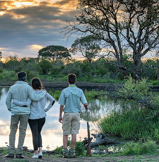A safari guide and two guests standing at the edge of a waterhole at sunset in Madikwe Game Reserve.