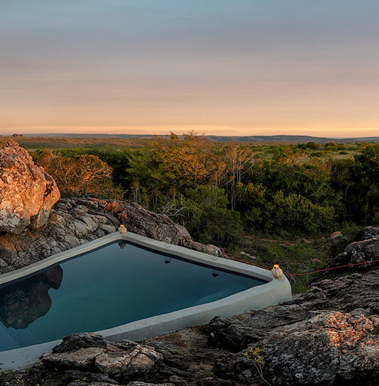 A private plunge pool built into a rocky ridge overlooking the open plains of Shamwari Private Game Reserve at sunset.