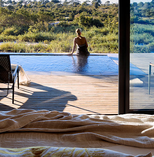 A woman sitting on the edge of a private plunge pool at a luxury lodge, overlooking the wilderness of Sabi Sand Private Game Reserve, South Africa.