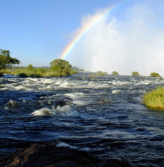 The Zambezi River flowing towards the edge of Victoria Falls, with the spray creating a rainbow, Livingstone, Zambia.