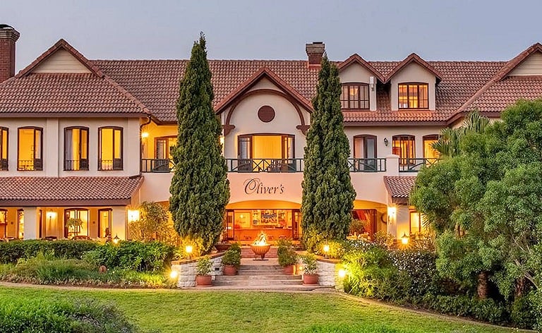 Two-storey hotel with a terracotta tile roof, lit windows and tall cypress trees, fronted by a lawn at dusk.