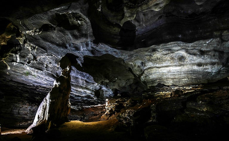Layered rock walls and a tall stalagmite lit by spotlights inside a dark cave chamber.