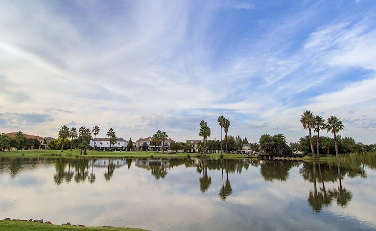 Palm trees and large houses reflected in a still lake beneath streaky clouds and blue sky.