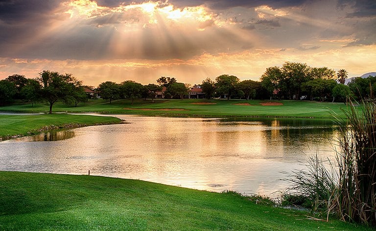 Sunbeams breaking through clouds over a water hazard, with fairways and red sand traps beyond.