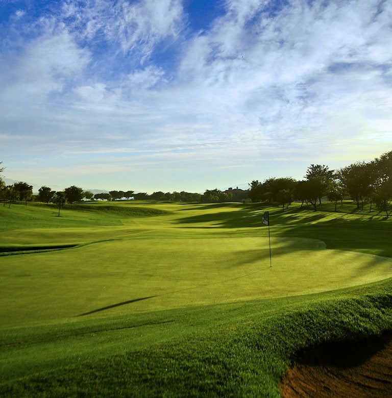 Flagstick on a sunlit putting green, with rolling fairways and trees in the distance under a blue sky.