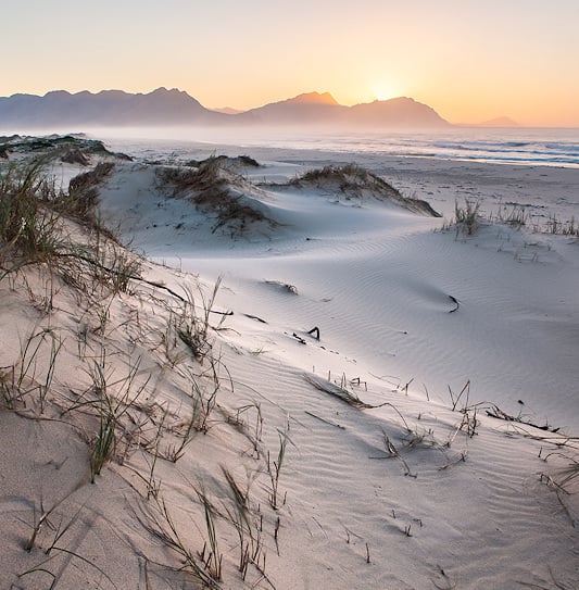 Grass-tufted sand dunes and a wide beach at sunset, with sea mist drifting below a mountain range.