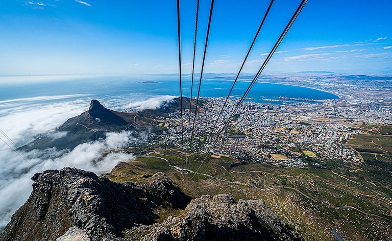 View along cable car wires over a pointed peak, city sprawl and a bay, with low cloud on the left.