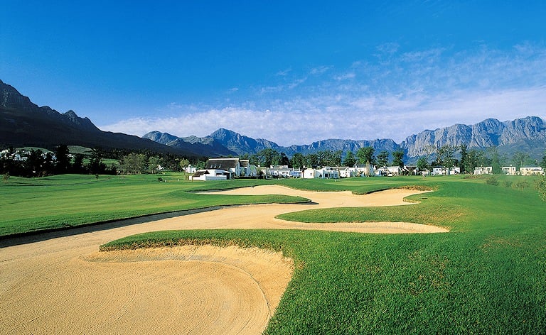 Wide sand traps cutting across a fairway, with white houses and rocky peaks behind.