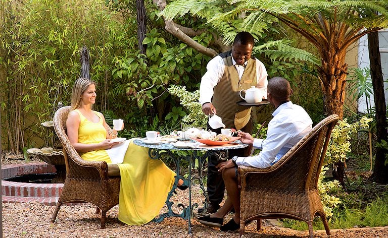 Waiter pouring tea for a couple seated in wicker chairs at a garden table among ferns.
