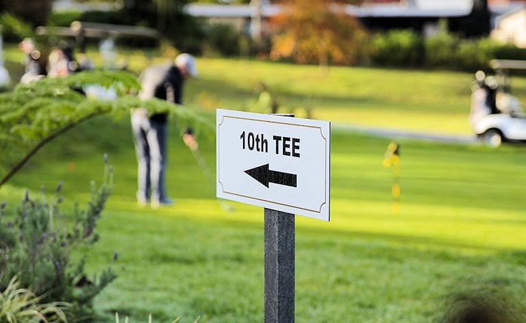 Sign with an arrow pointing to the 10th tee, with golfers on a green in the background.