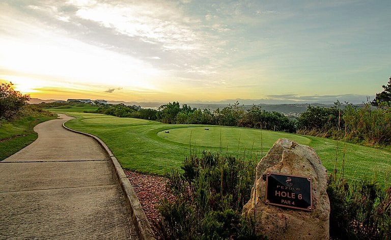 Curving cart path beside an engraved tee marker stone, with the fairway and distant hills in low golden light.
