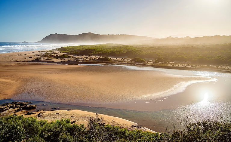 River mouth meandering across a sandy shore toward the ocean, with hazy headlands beyond.