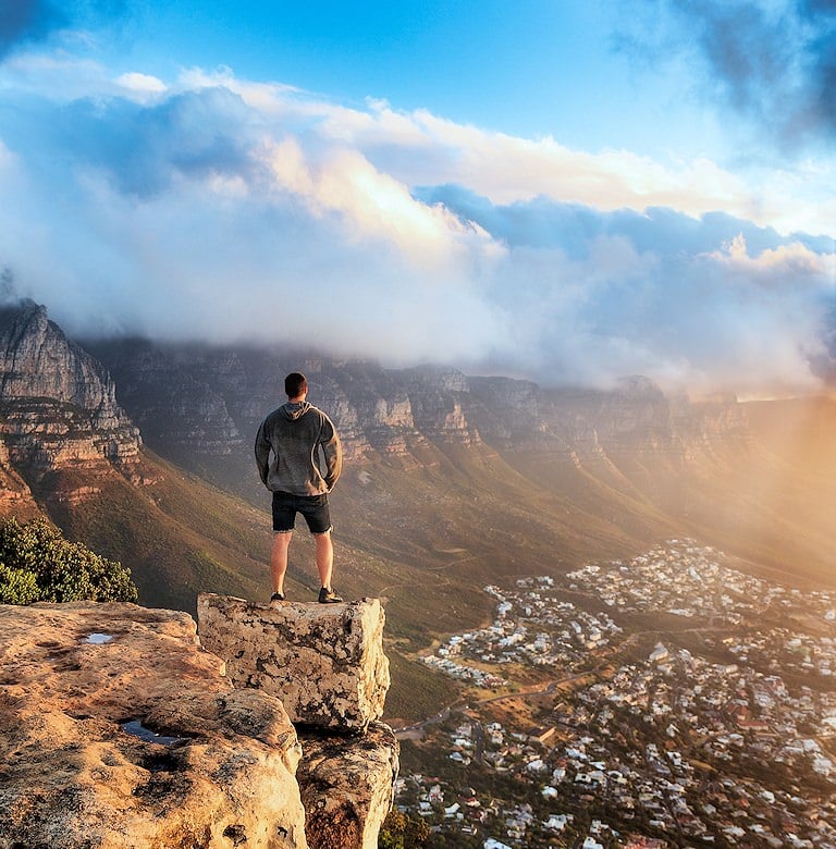 Hiker standing on a rock ledge looking over a town and a cloud-capped mountain range.