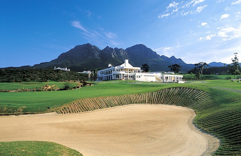 White clubhouse beyond a large raked bunker and fairway, with jagged mountains under a blue sky.
