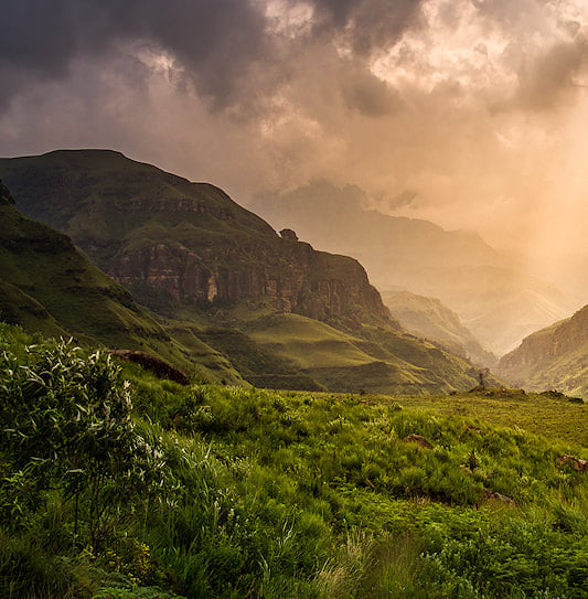 Sun rays breaking through storm clouds over a green Drakensberg valley flanked by sandstone cliffs and grassy slopes.