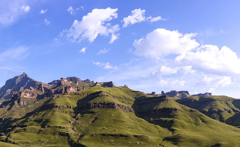 Grass-covered ridges rising to jagged rocky peaks under a blue sky with scattered white clouds.