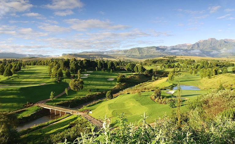 Fairways, bunkers and a footbridge over a stream, with a mountain escarpment on the horizon in morning light.