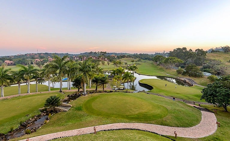 Elevated view over a putting green, palm trees and a winding pond on a resort golf course at dusk.