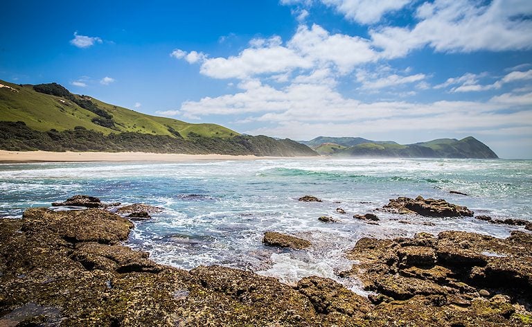 Waves washing over a rocky shoreline, with a sandy beach and green coastal hills in the distance.