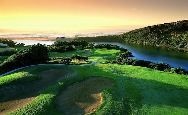 Sculpted bunkers on a coastal hole overlooking a lagoon, beach and ocean at sunset.