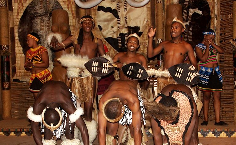 Dancers in traditional beaded dress and animal skins performing with cowhide shields.