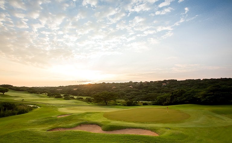 Flag on a green guarded by a long sand trap, with woodland and a hazy sky beyond.