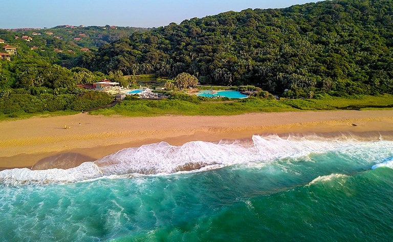 Aerial view of waves breaking on a wide beach below forested slopes and resort swimming pools.