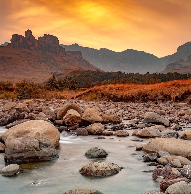 Shallow river flowing over smooth boulders beneath rugged cliffs and an orange evening sky.