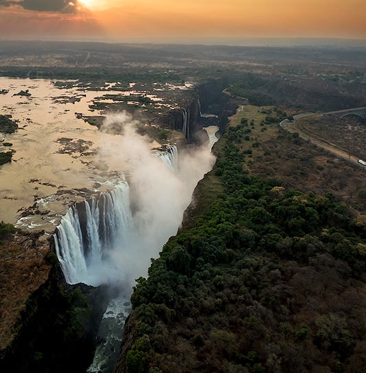Aerial view of Victoria Falls at sunrise, with the Zambezi River plunging into the gorge near the border bridge.