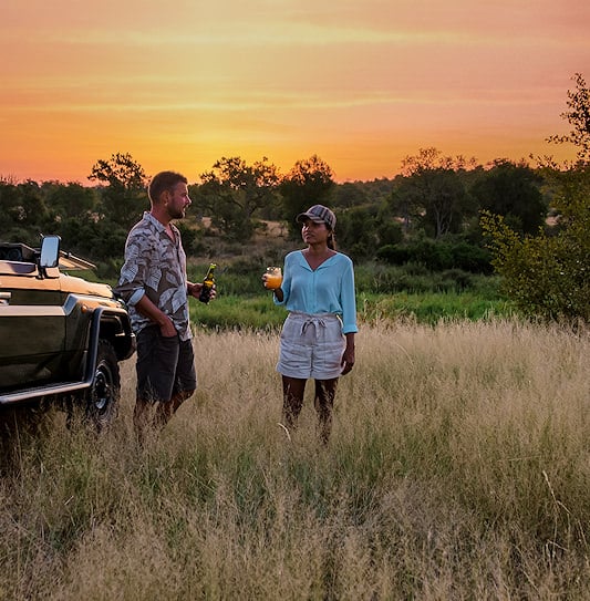 A man and a woman having sundowners next to an open-top safari vehicle in Kruger National Park.