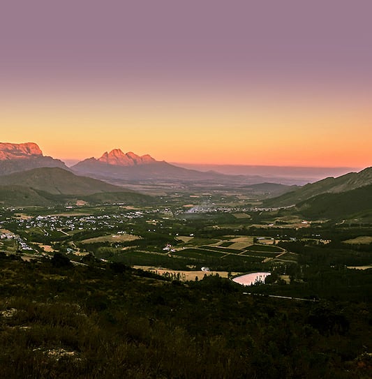 View of the Franschhoek Mountains and valley town from the pass under a purple and orange gradient dawn sky.