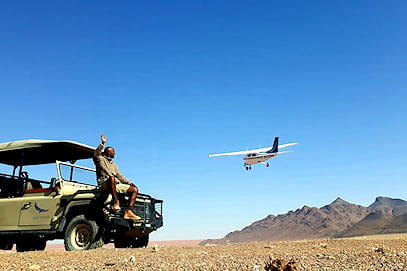 Safari guide sitting on a 4x4 vehicle waving to a low-flying light aircraft in the desert.