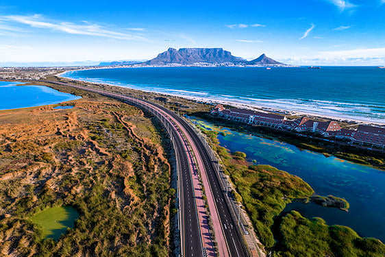 Aerial view of a coastal highway along the ocean with Table Mountain in the distance.