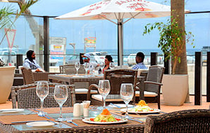 Diners sitting at outdoor restaurant tables under a parasol overlooking the ocean.