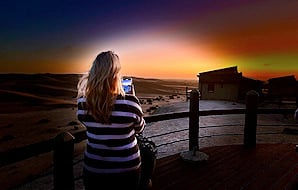 A tourist taking a photo of sand dunes with a smartphone from a wooden deck during sunset.