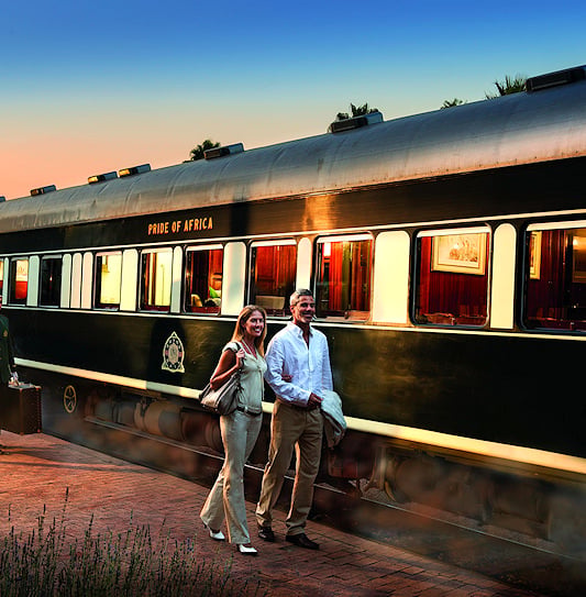 A couple walking next to a Rovos Rail train with a staff member carrying their luggage.
