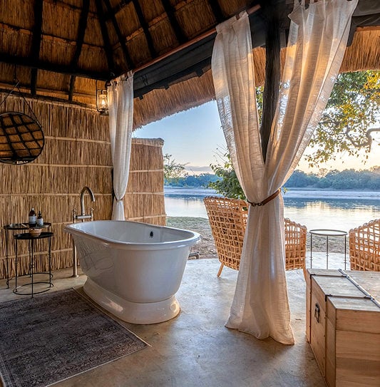 Open-plan bathroom featuring a freestanding tub overlooking a river sunset in South Luangwa, Zambia.