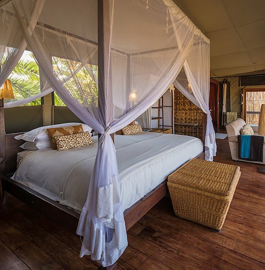 Tented suite bedroom with a canopy bed WITH white netting overlooking palm trees in the Busanga Plains, Zambia.