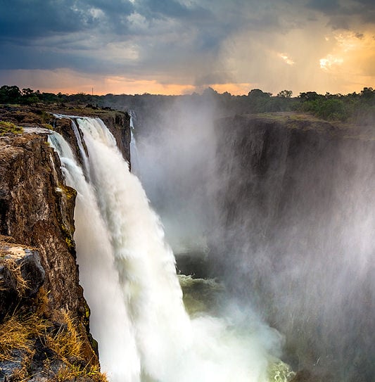Water plunging over the sheer cliffs of Mosi-oa-Tunya into a misty gorge under a dramatic sunset sky.