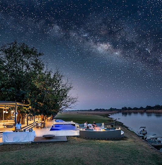Visitors socializing around a fire pit at a lodge under a starry night sky in South Luangwa National Park, Zambia.
