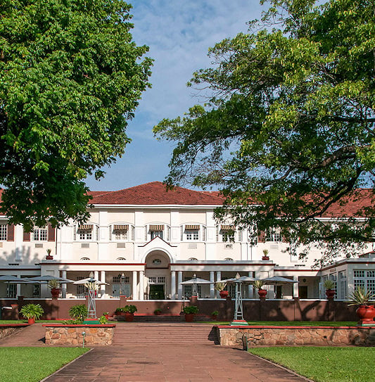 White colonial building with red tiled roof framed by large shade trees in Victoria Falls.